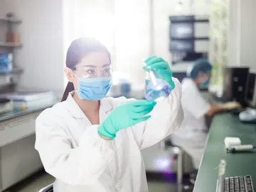 A woman in white lab coat and mask holding up a glass.